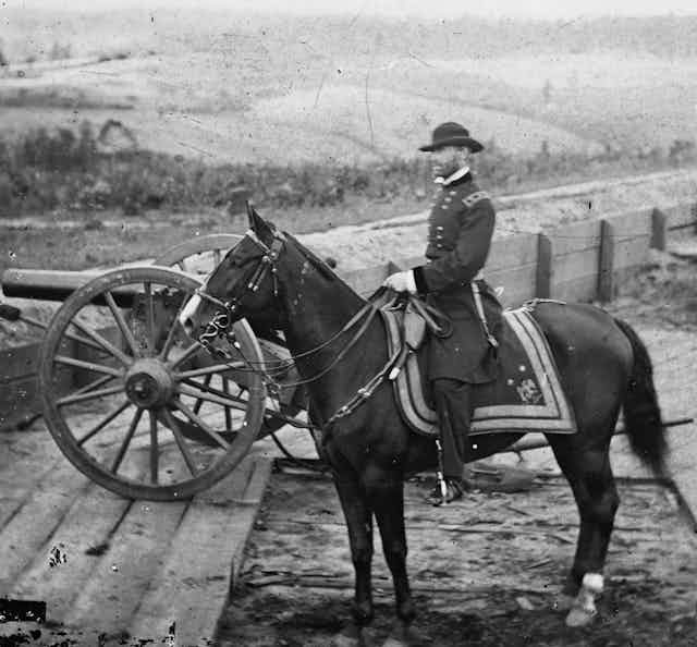 A black and white image of a man in a military uniform sitting on a horse, behind some wooden fortifications and a cannon