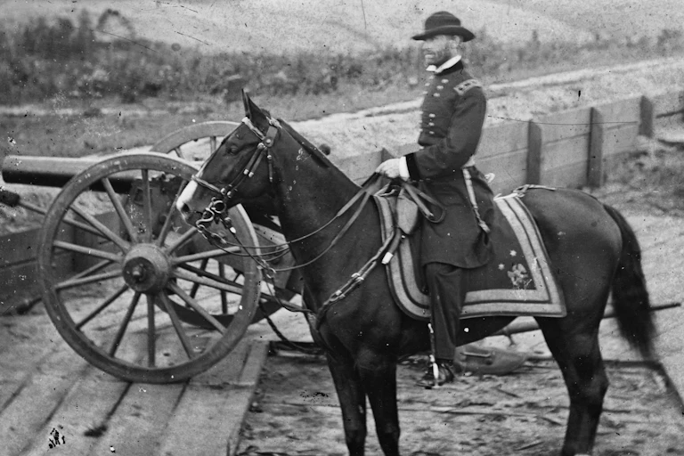 A black and white image of a man in a military uniform sitting on a horse, behind some wooden fortifications and a cannon