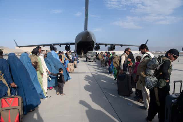 Two rows of people are seen awaiting to board a large military plane on a sunny day