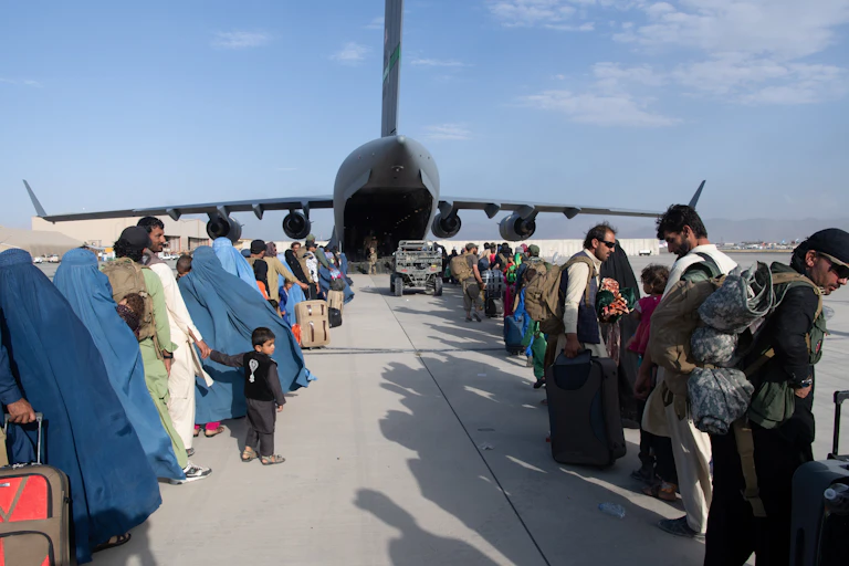 Two rows of people are seen awaiting to board a large military plane on a sunny day