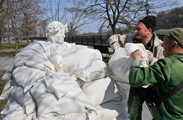 A statue of a man wearing a leaf crown is surrounded by sandbags applied by two workers.
