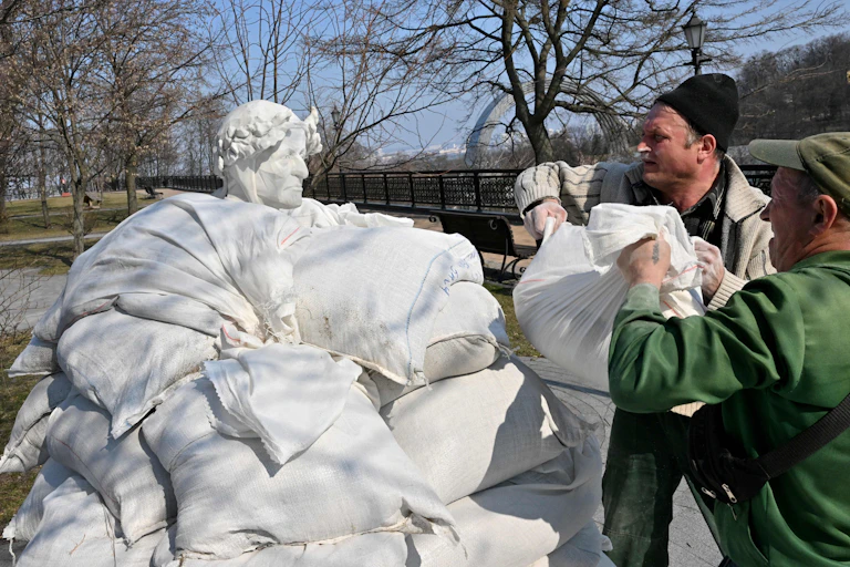 A statue of a man wearing a leaf crown is surrounded by sandbags applied by two workers.