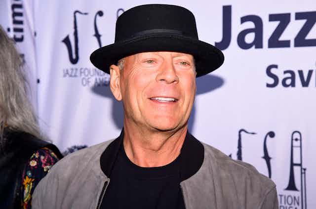 The actor Bruce Willis sporting a black pork pie-type hat stands in front of a banner for a jazz festival