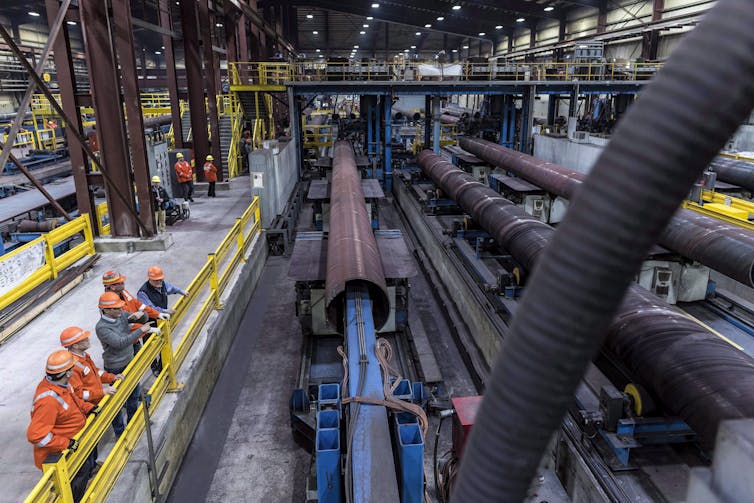 Workers in construction uniforms stand at a railing overlooking a steel factory