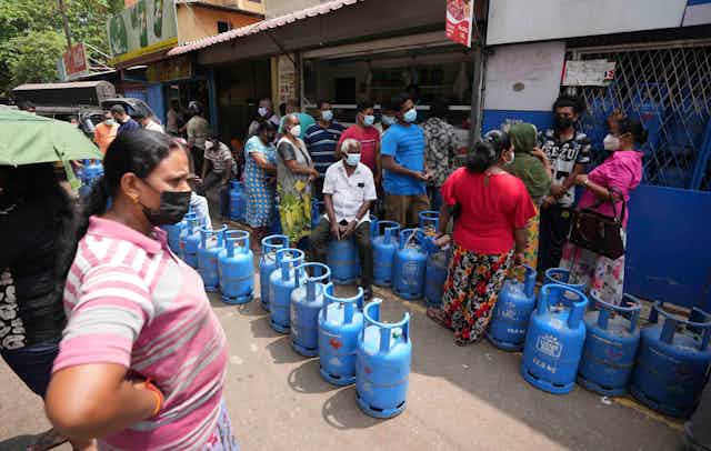 Empty blue cylinders of gas line the street as a variety of men and women in colorful shirts and masks stand nearby with stores in the background