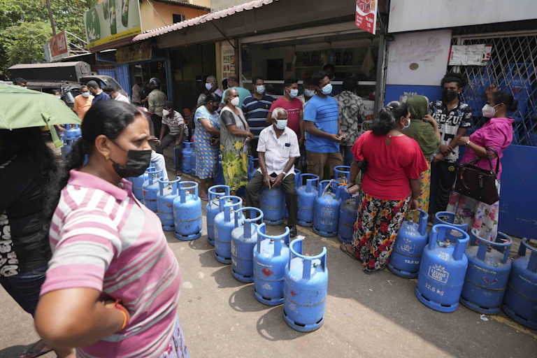 Empty blue cylinders of gas line the street as a variety of men and women in colorful shirts and masks stand nearby with stores in the background