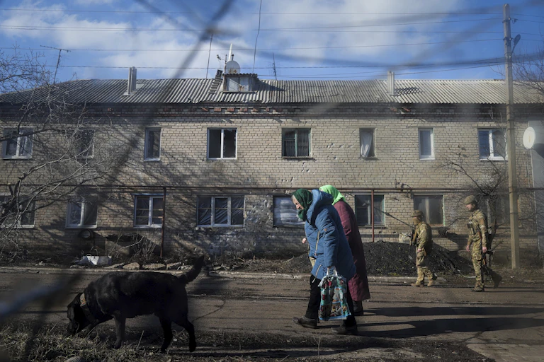 two soldiers patrol a street as elderly women and a dog walk past a house