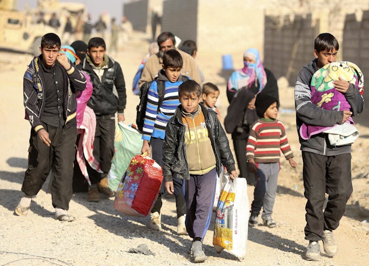 A group of teens and children carrying their belongings leave a desert village.