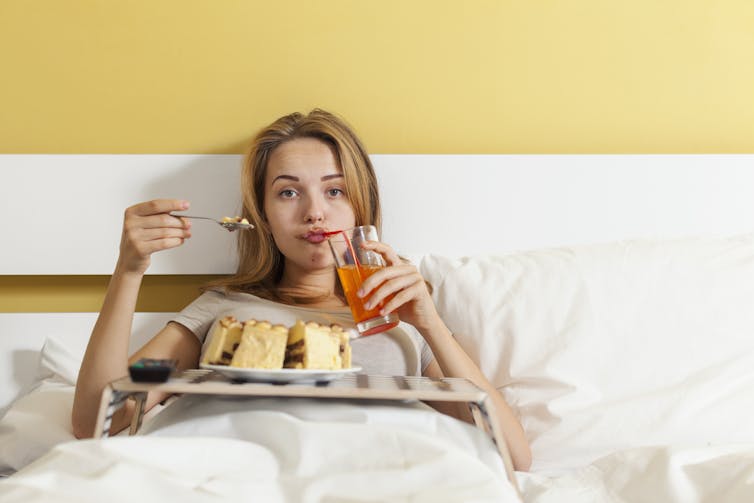 A young woman sitting in bed eating cake and drinking juice with a bored expression on her face.