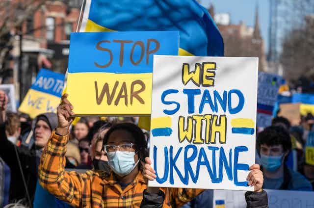 A man wearing a surgical mask, glasses and a plaid shirt holds up two signs during a protest. One says 'stop war' and the other says 'we stand with Ukraine.'