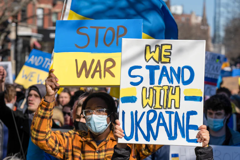 A man wearing a surgical mask, glasses and a plaid shirt holds up two signs during a protest. One says 'stop war' and the other says 'we stand with Ukraine.'