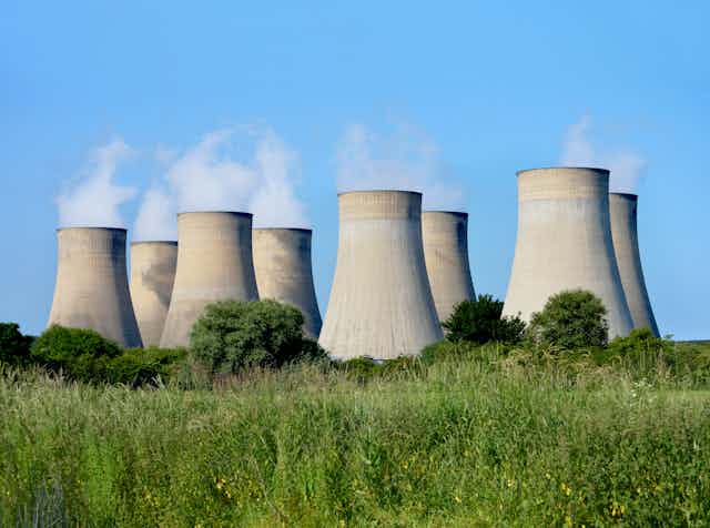 Eight cooling towers releasing steam against a blue sky with a treeline in the foreground.