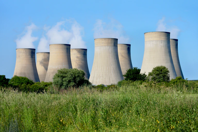 Eight cooling towers releasing steam against a blue sky with a treeline in the foreground.