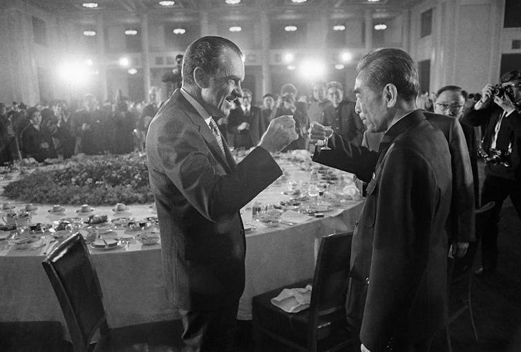 Two smiling older men toast each other as they stand in the front of a banquet table and are watched by a crowd of people.