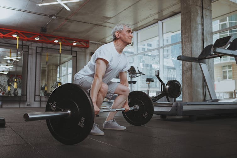 An older man perform a deadlift using a barbell in a gym.