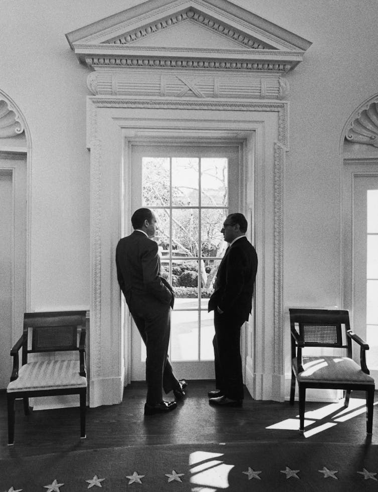 Two men in suits talking to each other in a large, elegant room with high ceilings, standing next to a window.