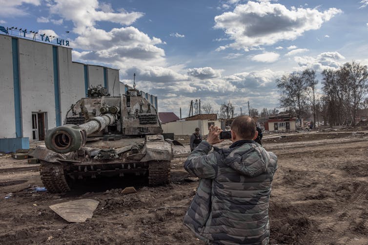 A man takes a photograph of a tank outside buildings.