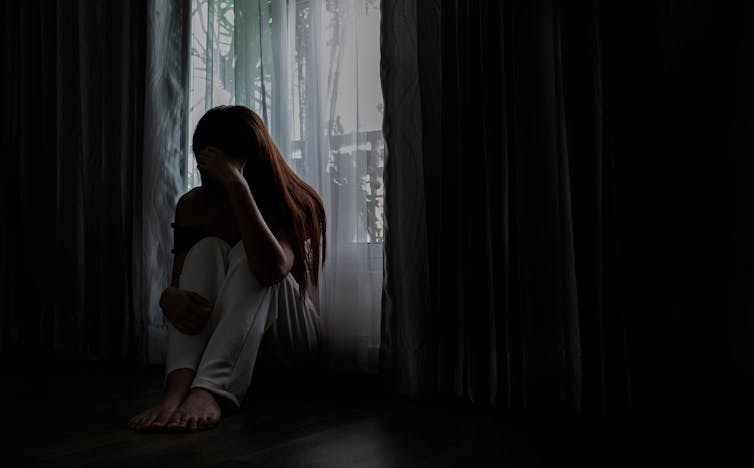 A woman sitting in shadow on the floor in a bedroom by the window, with her head in her hands