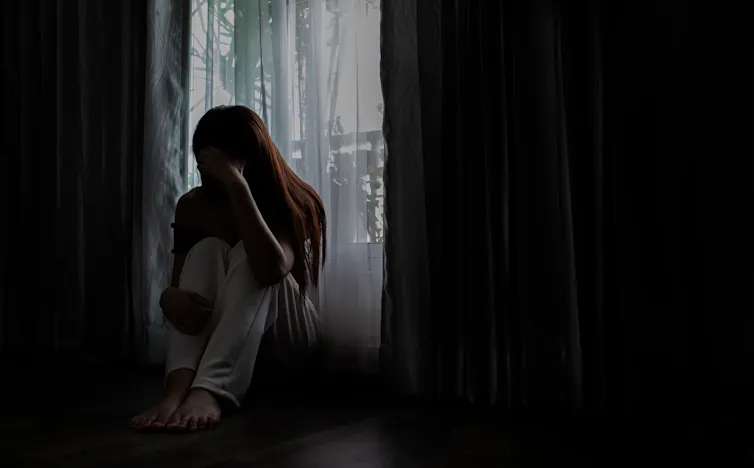 A woman sitting in shadow on the floor in a bedroom by the window, with her head in her hands