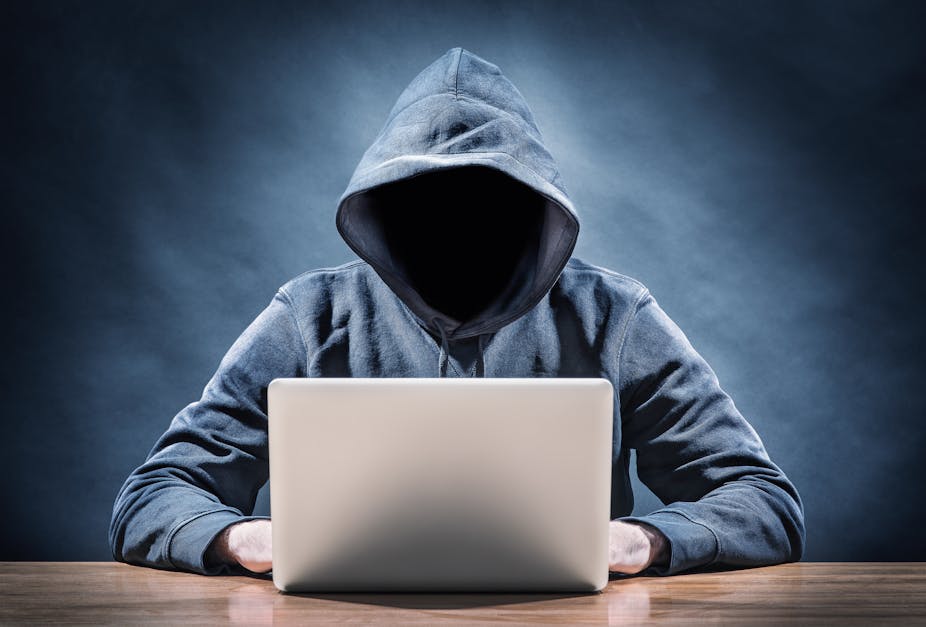 A person in a hoodie, with their face hidden by a dark shadow, uses a laptop computer