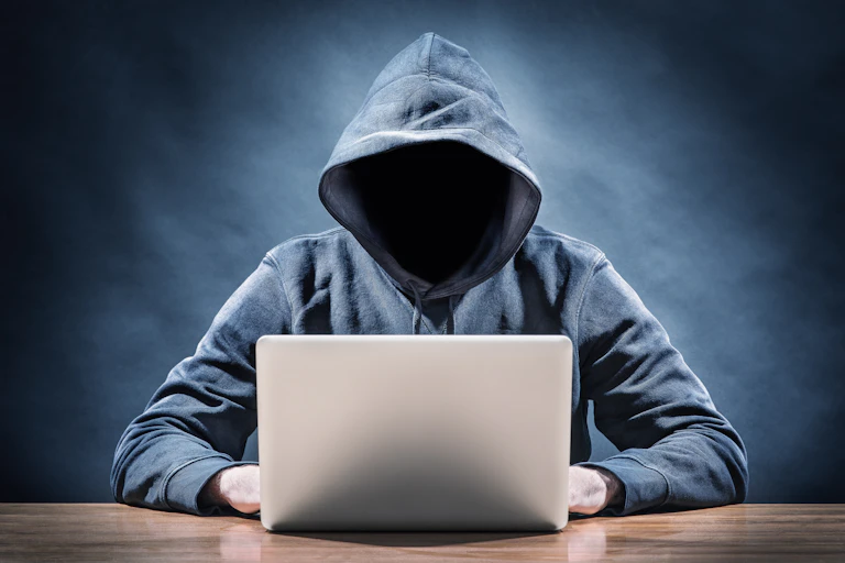 A person in a hoodie, with their face hidden by a dark shadow, uses a laptop computer