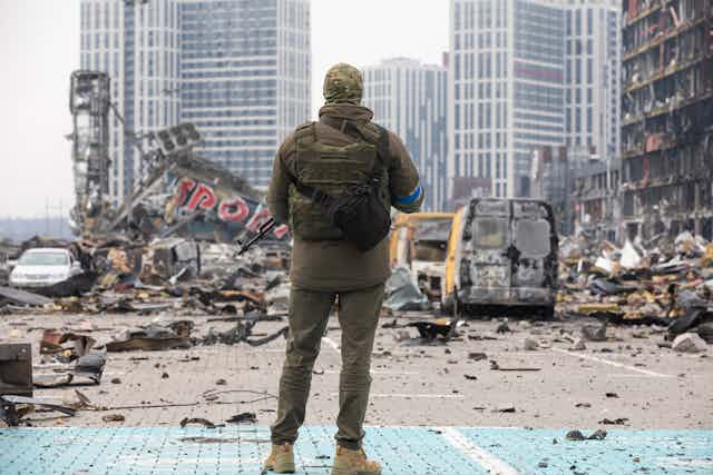 The back of a soldier wearing camouflage is shown looking at a destroyed building and vans on a grey day
