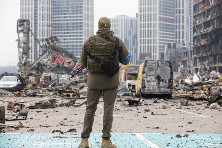 The back of a soldier wearing camouflage is shown looking at a destroyed building and vans on a grey day