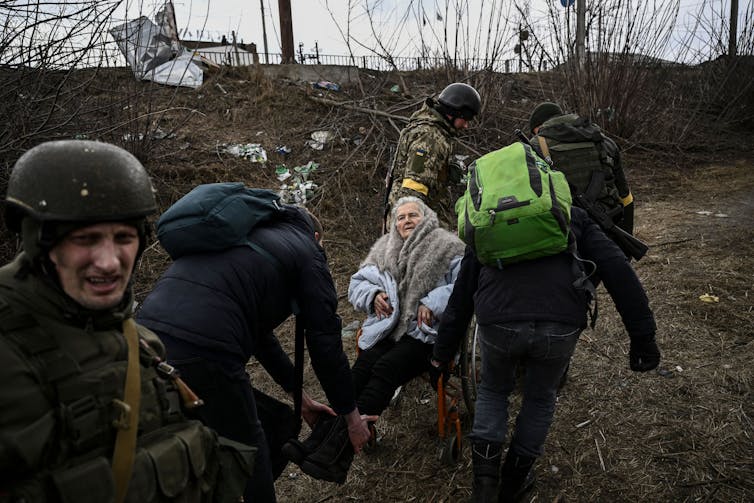 An elderly woman is seated in a wheelchair, carried across dirt by five men, some wearing army uniforms