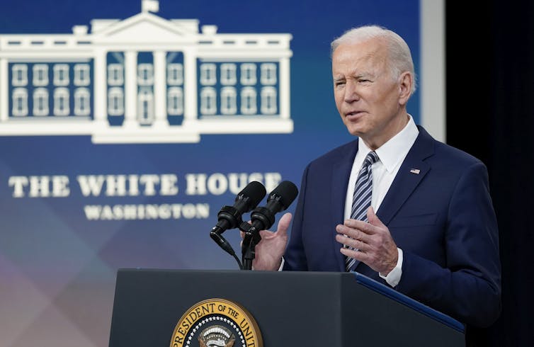 Elderly white guy wearing a blue suit stands in front of a podium with the seal of the US president and gesticulates with both hands as a logo of the white house is displayed in the background