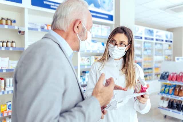 A female pharmacist wearing a mask holds medicine packages and explains instructions to an elderly person.