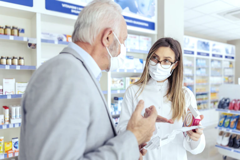 A female pharmacist wearing a mask holds medicine packages and explains instructions to an elderly person.