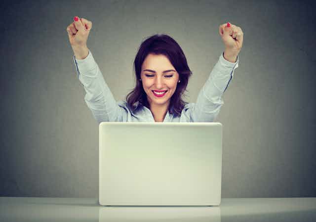 A woman smiles and raises her arms in triumph while looking at her laptop.