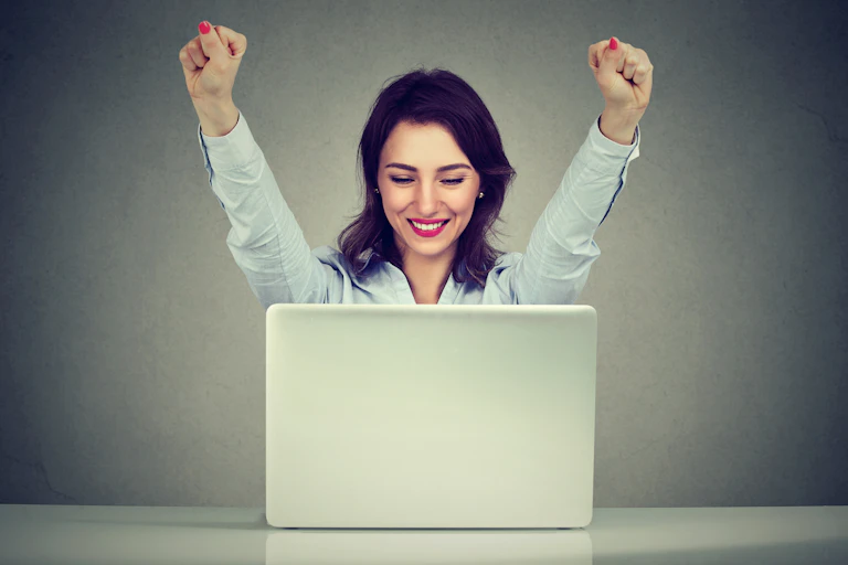 A woman smiles and raises her arms in triumph while looking at her laptop.