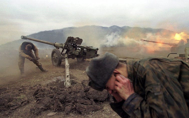 A soldier bows his head and plugs his ears as artillery is fired.