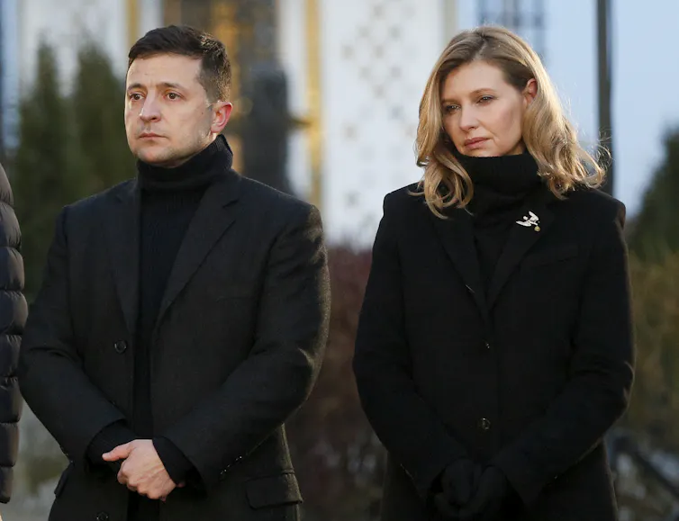 A dark-haired man stands next to a blonde woman. Both are dressed in black.