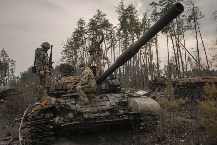 A soldier standing on a destroyed tank takes a selfie on his smartphone.