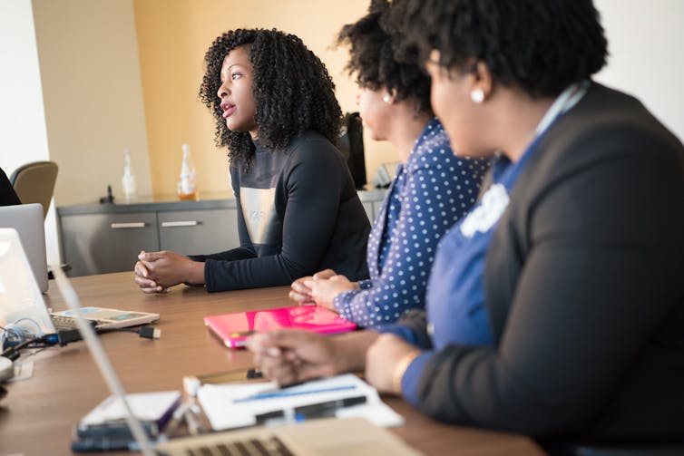 Three people sitting at a table with laptops; two are watching the third as she speaks