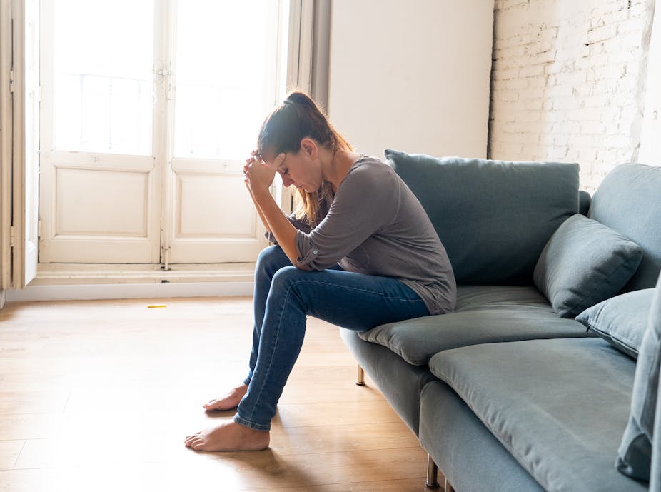 Young woman sitting on a couch alone holds her head in her hands looking sad.