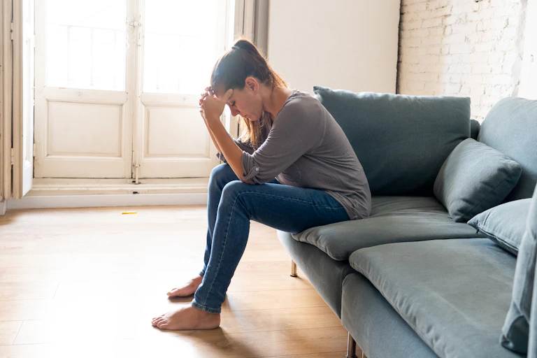 Young woman sitting on a couch alone holds her head in her hands looking sad.