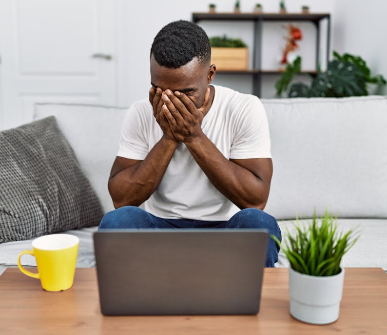 A young man sits in front of a laptop computer with his hands covering his face