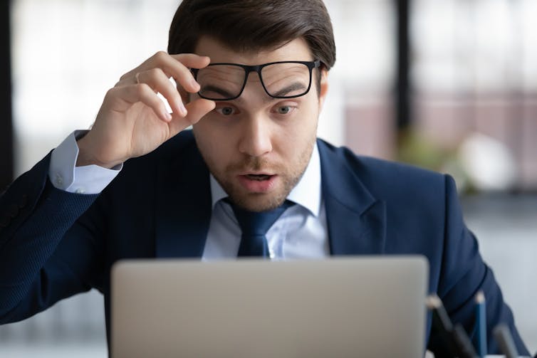 A man in a suit lifts his glasses and looks shocked while looking at a computer.