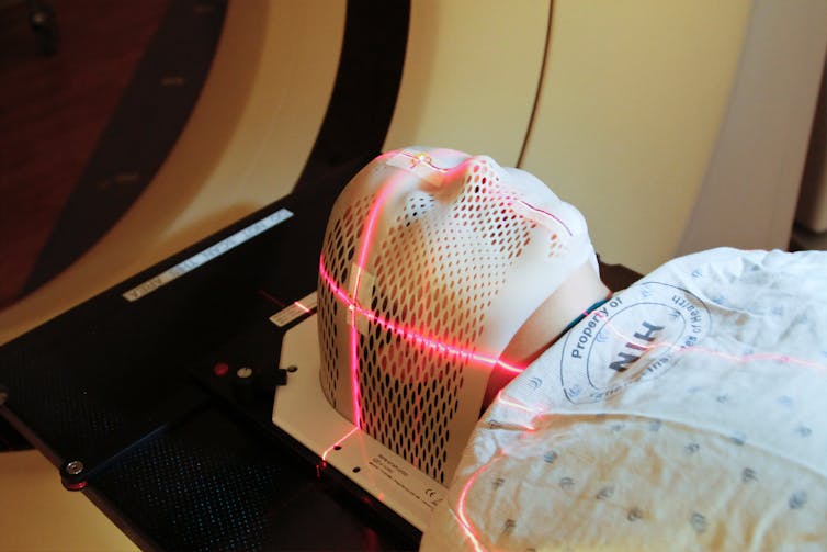 A person lying down wearing a hospital gown with a mesh mask over their head, with red lines of light