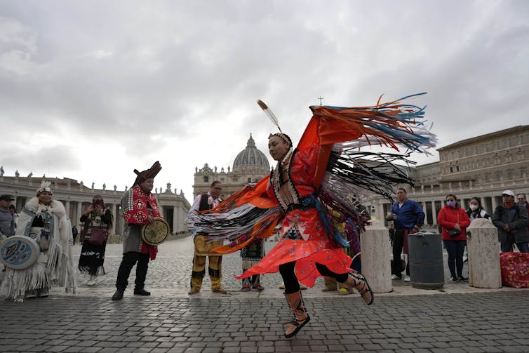 A dancer in regalia wearing orange is seen in a public square in front of some people holding drums.