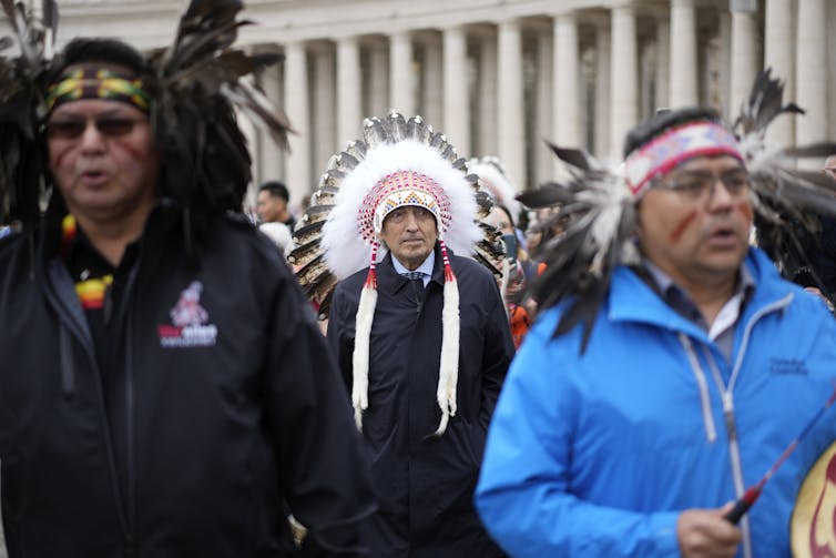 A man in suit and tie and feather regalia seen standing between two men, one holding a drum.