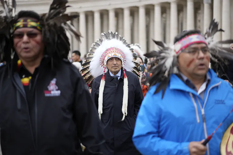 A man in suit and tie and feather regalia seen standing between two men, one holding a drum.