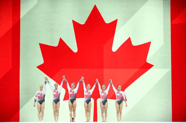 A group of gymnasts stand next to eachother in front of the Canadian flag