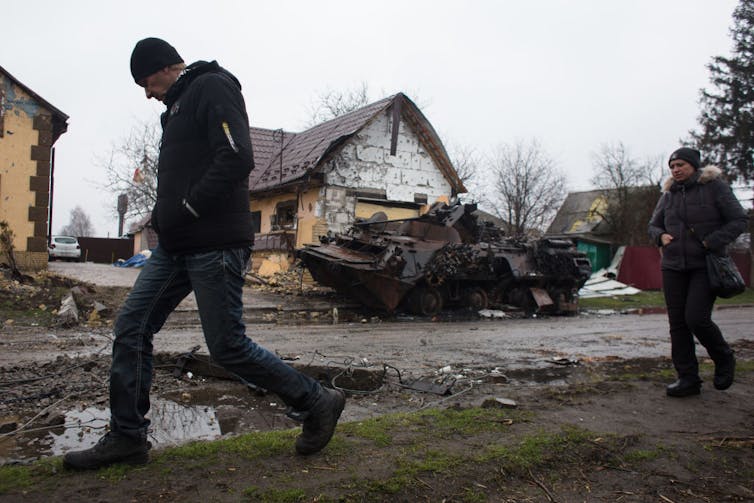 Two people wearing winter jackets walk past a burned tank on a muddy road in front of what could be residential or farm buildings