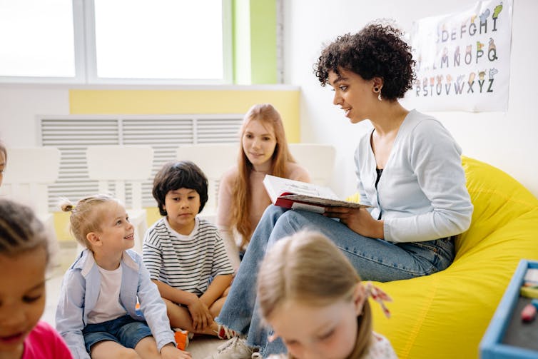 A woman is seen holding a book reading to a group of young children.