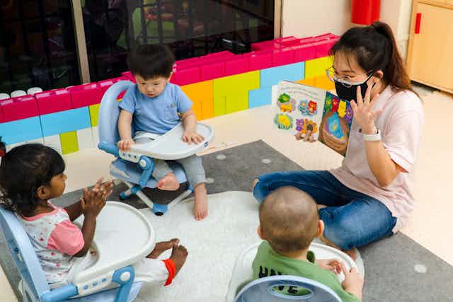A woman seen in a face mask holding a children's book reading to children in chairs.