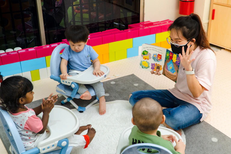 A woman seen in a face mask holding a children's book reading to children in chairs.